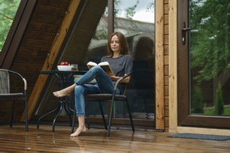 Barefoot woman sits on a chair on a wet wooden deck of tiny house after the rain. She is reading a