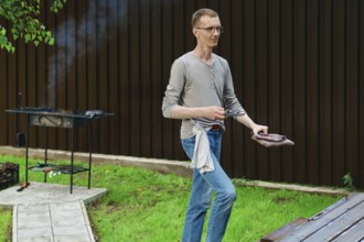 A man stands in a backyard holding a tray with meat while grilling on a charcoal grill. Smoke rises