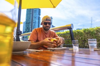 Tourist wearing sunglasses and a baseball cap enjoys a burger and beer on an outdoor patio