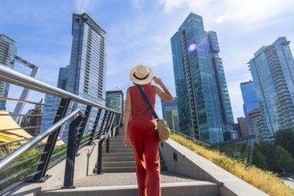 Female tourist wearing a straw hat and holding her hat is walking up stairs with modern glass