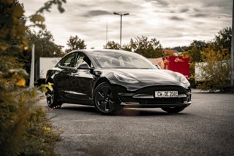 Black car on asphalt surface with cloudy sky and autumn vegetation, Deer E-Carsharing, Tesla Model