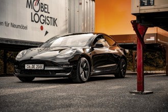 Black car in front of furniture trailers in bright sunlight on a road surface, Deer E-Carsharing,