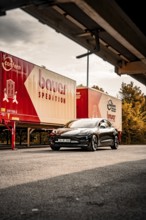 Black car in front of trucking trailers on a road with cloudy sky and trees, Deer E-Carsharing,