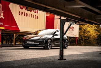 Black car parked next to freight forwarding trailers under a bridge-like structure, autumn trees in