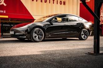 Black car on a road in front of a bright wall with red details, Deer E-Carsharing, Tesla Model 3,