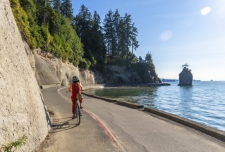 Tourist wearing helmet and red clothes enjoys cycling in stanley park seawall with siwash rock and