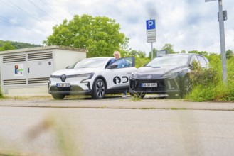 Two electric cars are parked in a parking lot, and a man is standing next to them. Cloudy sky and