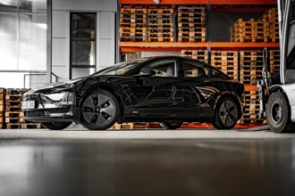 Black car in a warehouse next to wooden pallets and machines, Deer e-Carsharing, Tesla Model 3,