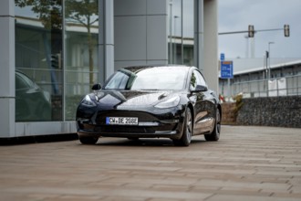 Black car in front of a modern building with wet road and cloudy sky, Deer E-Carsharing, Tesla