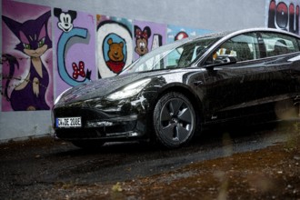 Black car in front of a rain-covered road and colorful graffiti wall, Deer E-Carsharing, Tesla