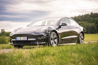 Black car on a country road surrounded by green fields under cloudy sky, Deer E-Carsharing, Tesla