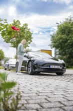 Person standing next to black car on paved road under blue sky with clouds, Deer E-Carsharing,