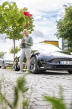 Woman using a tablet next to a parked black car outdoors, Deer E- car sharing, Tesla Model 3, Calw,