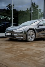 Black car in front of a glass building on rain-wet ground, Deer E-Carsharing, Tesla Model 3, Calw,