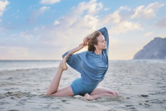 A woman practices Eka Pada Rajakapotasana on the sand during sunset, focusing on her yoga pose with