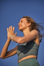 A woman practices yoga on the beach during sunset, holding her hands in Namaskar Mudra while
