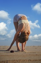 A woman performs Uttanasana on a sandy beach during sunset while clouds fill the sky. She leans