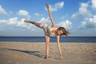 A woman practices Ardha Chandrasana Half Moon Pose on sandy beach while balancing on one leg and
