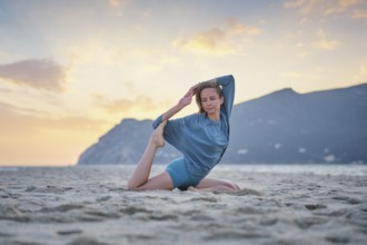 A woman performs Eka Pada Rajakapotasana on a sandy beach at sunset. The warm sky adds to the