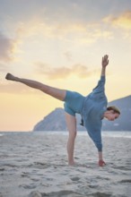 A woman does Ardha Chandrasana Half Moon Pose on the sand during sunset. She balances on one leg