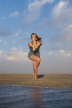 A woman stands on a sandy beach performing Eagle Pose with wrapped legs and crossed arms during