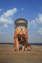A woman does Uttanasana on a sandy beach during sunset, focusing on her breath and stretching her