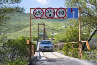 A car crosses a suspension bridge, accompanied by road signs with width and height restrictions