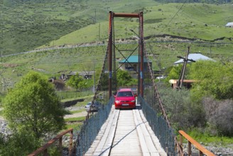 A red car crosses a narrow suspension bridge in a green, hilly landscape with houses in the