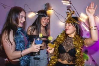 Three young women smiling, laughing, and drinking colorful cocktails while having fun at a festive