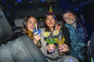 Three smiling young women wearing festive hats and tinsel, holding colorful cocktails, enjoying a