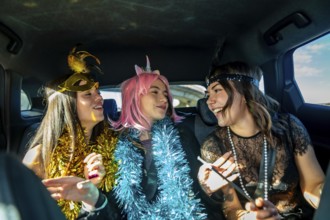 Three young women laugh and celebrate in a car during a colorful costume party, wearing masks, wigs