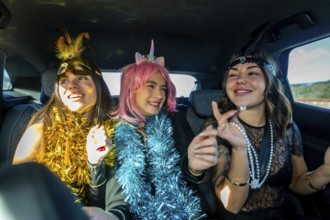 Three young women laughing and celebrating in a car back seat, wearing colorful tinsel boas and