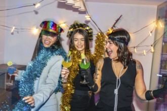 Three young diverse women smiling, laughing, and raising colorful cocktails, wearing festive