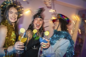 Diverse women in festive party hats and tinsel clink colorful lime garnished cocktails, laughing