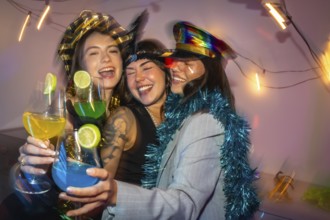 Three happy young women in festive costumes laughing and cheering with vibrant cocktails, enjoying