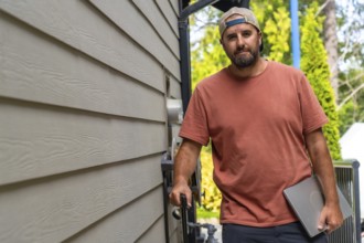 Maintenance worker walking alongside a residential building, carrying a laptop and various tools,