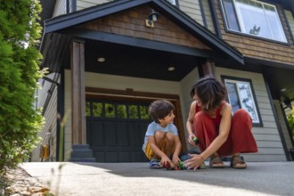 Mother and her young son are happily playing with toy cars on the driveway of their modern two
