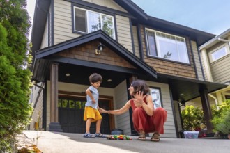 Mother interacts with her young son, playing with toy cars on the driveway of their modern suburban