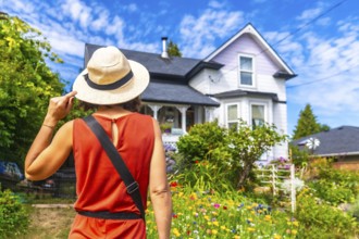 Female tourist wearing a straw hat, holding it with one hand, admiring a beautiful blooming garden