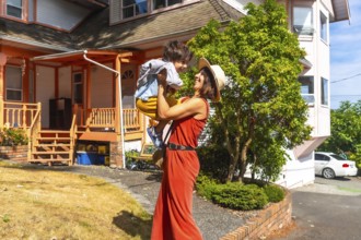 Happy mother wearing a straw hat and an orange jumpsuit lifting up her cheerful son in the yard of
