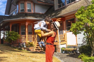 Happy mother lifting her son in the air, having fun in the yard of their colorful house in
