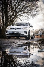 A white car with a reflected image is standing on a road in front of winter trees, VW ID4 electric