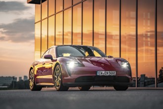 Red sports car in front of a building with glass façade at sunset, Porsche Taycan, Deer Carsharing,