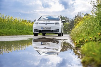 A white electric car on a country road, reflected in a puddle, surrounded by yellow flowers and