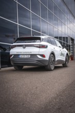 White car in front of glass façade on paved road, VW ID4 electric car, Deer Carsharing, Calw,