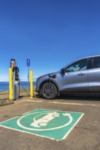 Modern electric car charging at a seaside station on a bright sunny day, highlighting the