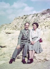 Portrait of smartly dressed man and woman, couple sitting on rocks at the seaside c 1956