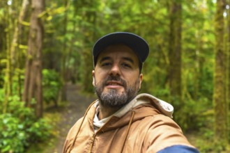 Tourist in a cap and light brown jacket taking a selfie while walking along a lush rainforest trail