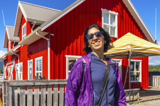 Happy tourist wearing sunglasses and purple jacket smiling in front of a vibrant red building in