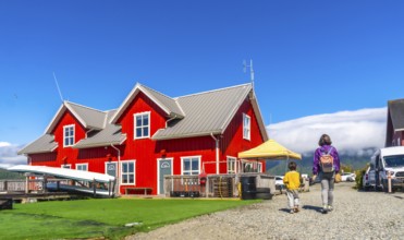 Mother and son walking on gravel path near vibrant red wooden building with canoes, enjoying summer